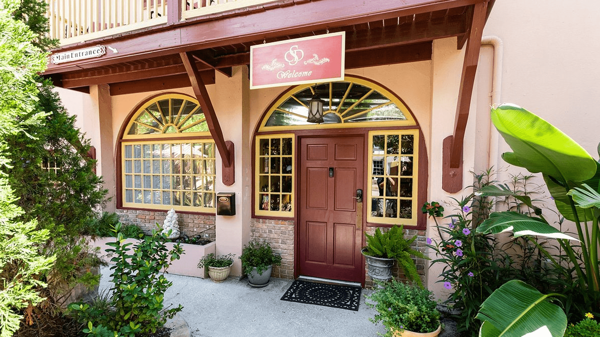 The entrance to Casa de Solana Bed and Breakfast, featuring a red door with yellow trim, arched windows, and a hanging sign that says "Welcome." There are potted plants and shrubs surrounding the building.