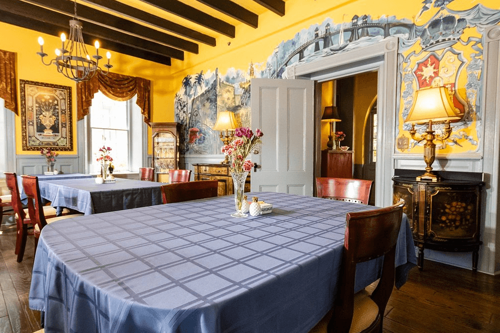 A dining room with yellow walls, a wooden ceiling, and a large mural depicting a bridge and a castle. There are several tables set with blue tablecloths and a chandelier hanging from the ceiling.