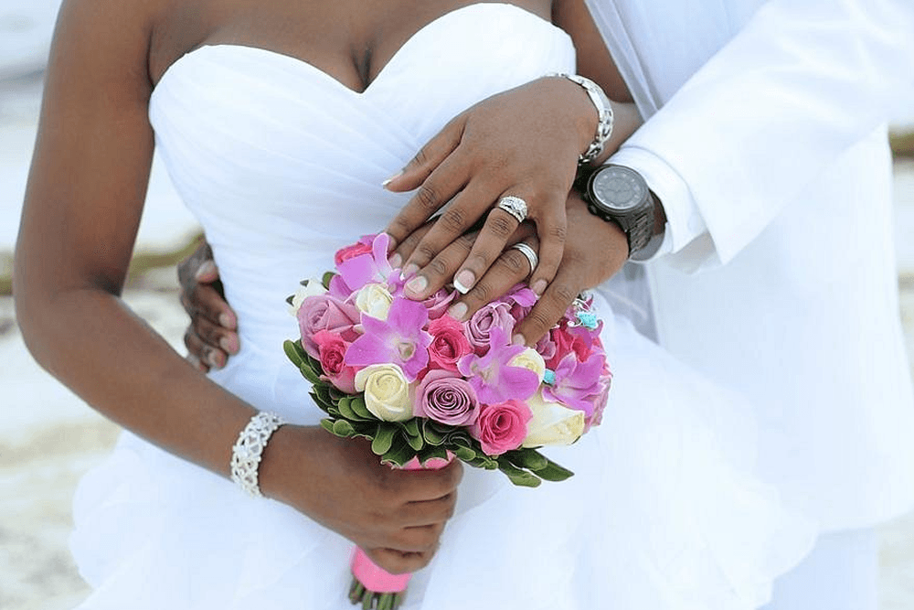 Man and woman in wedding clothes showing off rings over bouquet of pink and white flowers