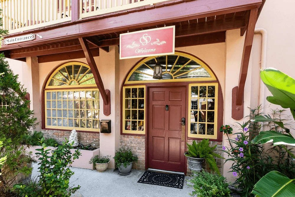 The entrance to Casa de Solana Bed and Breakfast, featuring a red door with yellow trim, arched windows, and a hanging sign that says "Welcome." There are potted plants and shrubs surrounding the building.