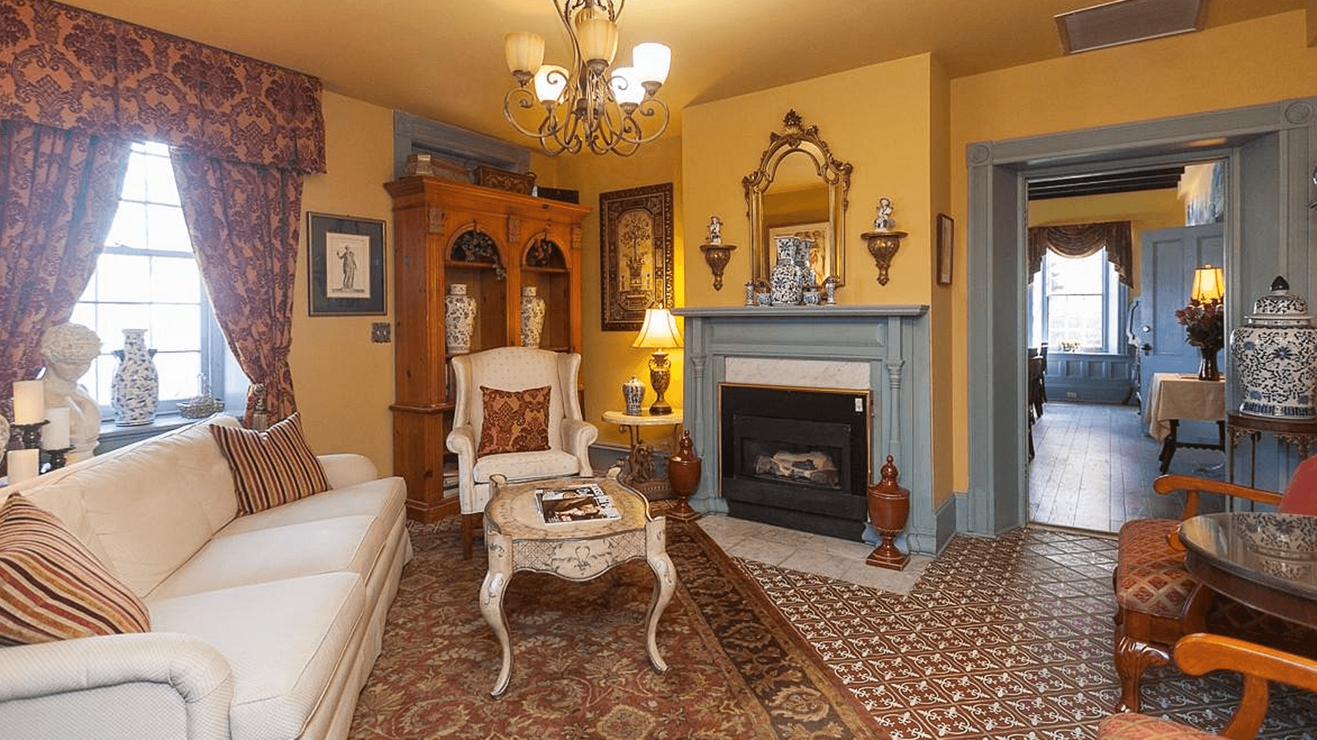 A cozy living room with a fireplace, a white sofa, and a patterned rug. There is a wooden bookcase filled with decorative items and a chandelier hanging from the ceiling.