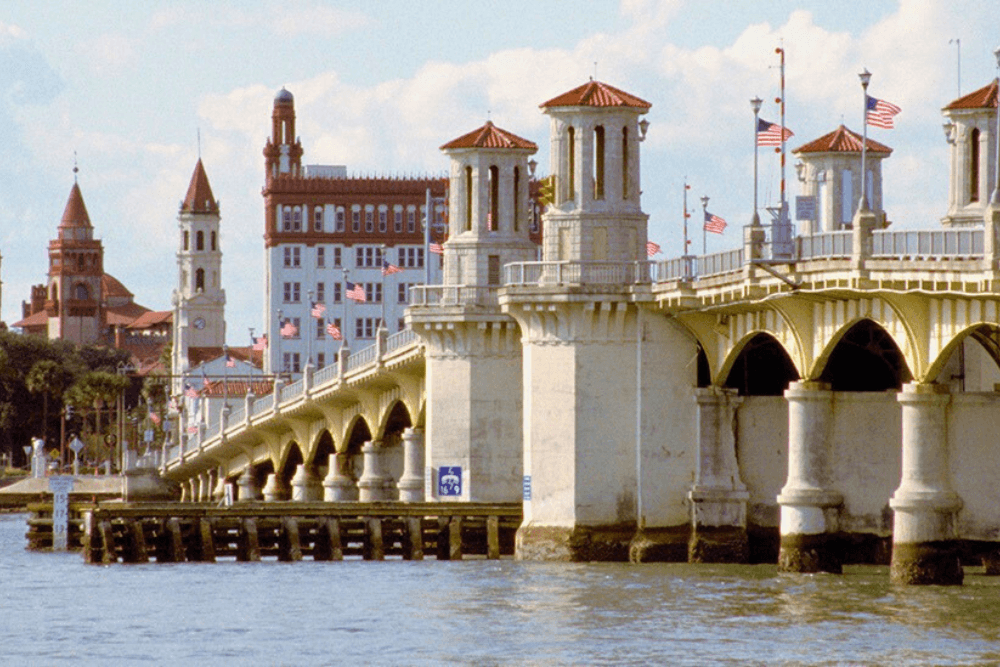 A view of the Bridge of Lions in St. Augustine, Florida. The bridge is a historic concrete arch bridge with decorative towers and railings. There are buildings and flags visible in the background.