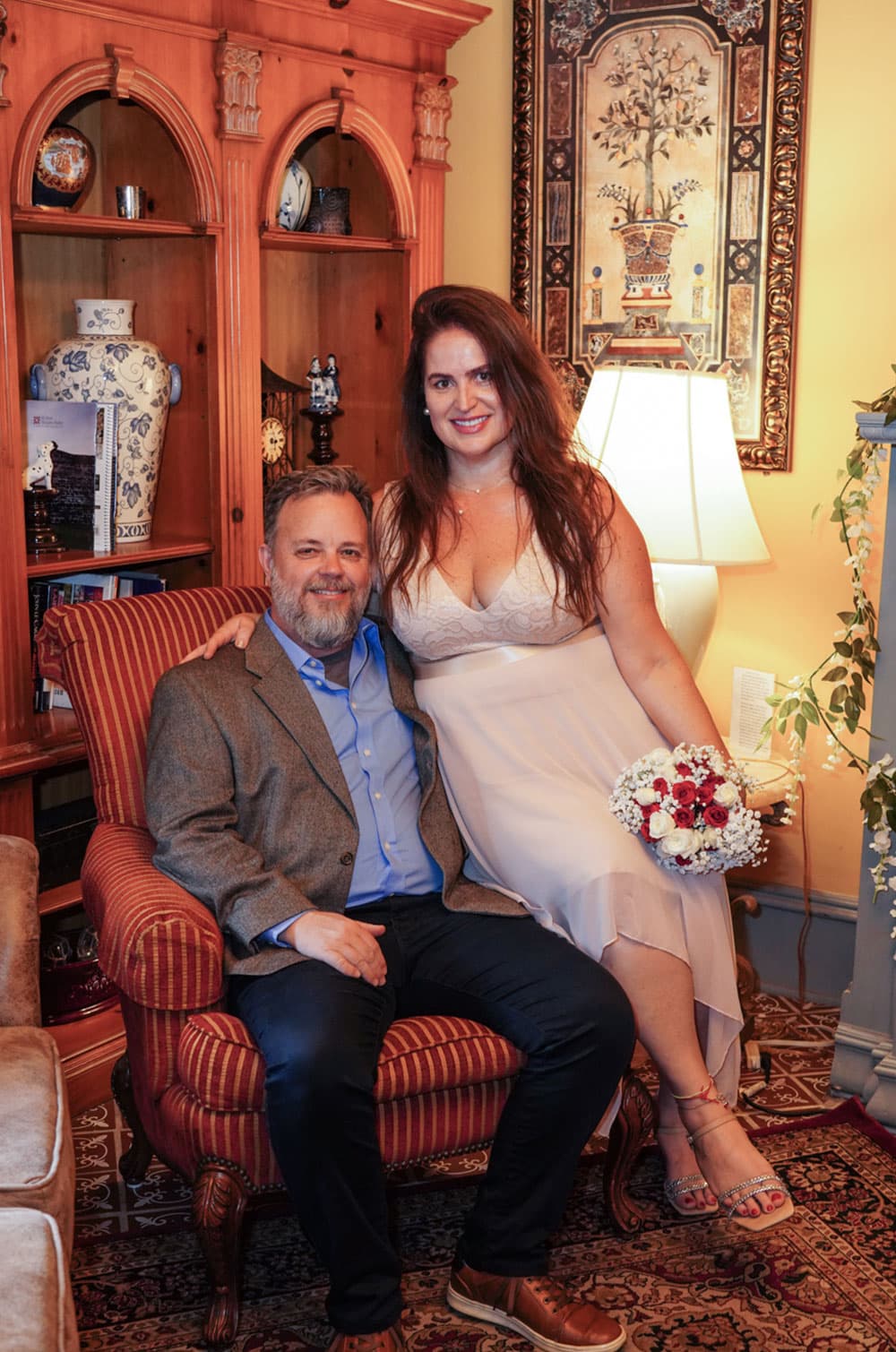A couple smiles while seated together in a cozy, warmly lit room, with the woman holding a bouquet.