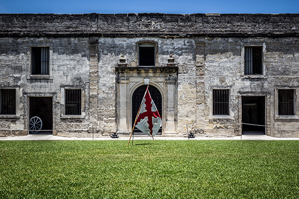 Gray stone Castillo de San Marcos with red and white flag in center doorway, green lawn in foreground