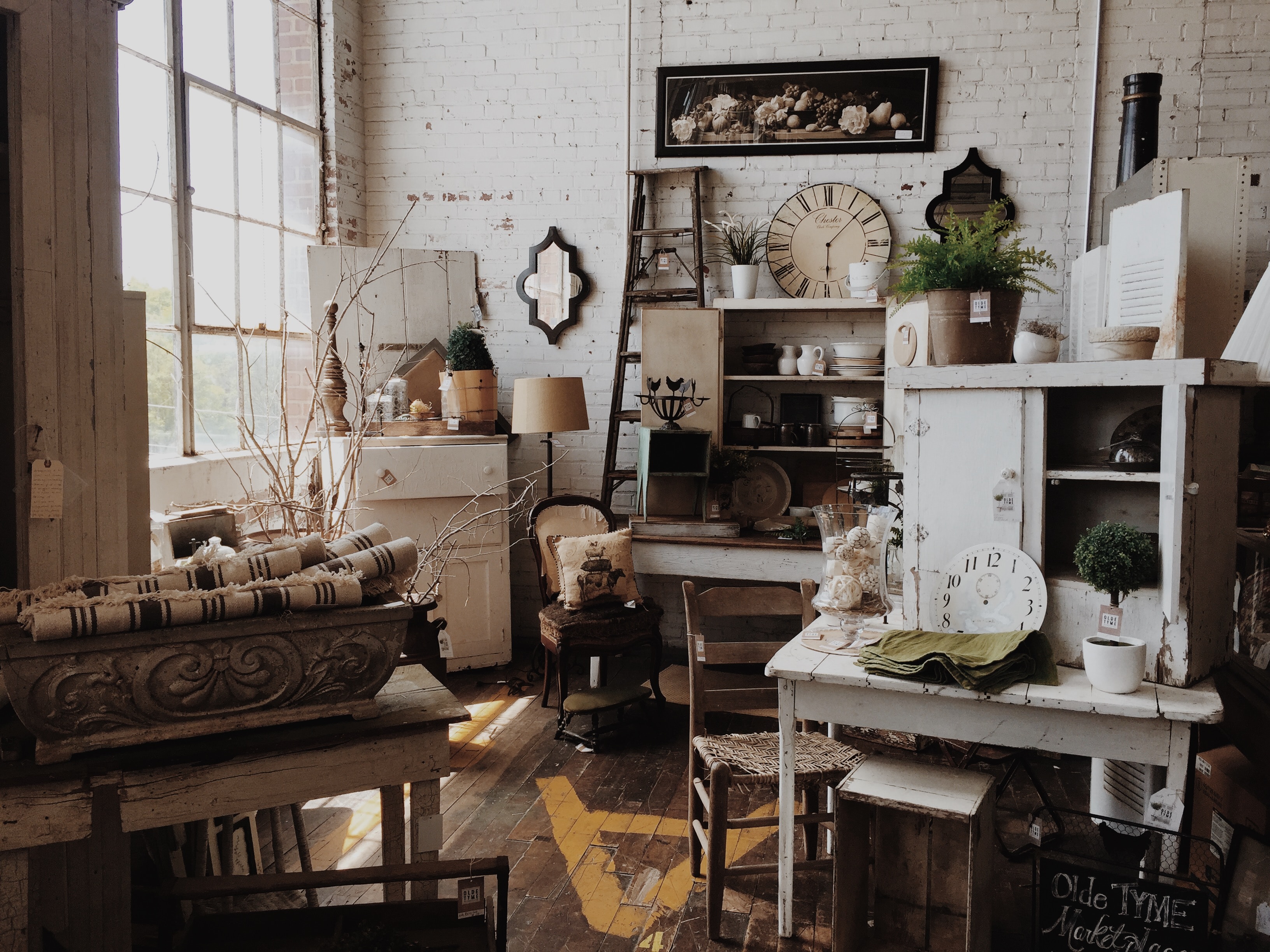 A naturally lit room full of white and brown antiques, including furniture, clocks, lamps, a ladder, and pottery.