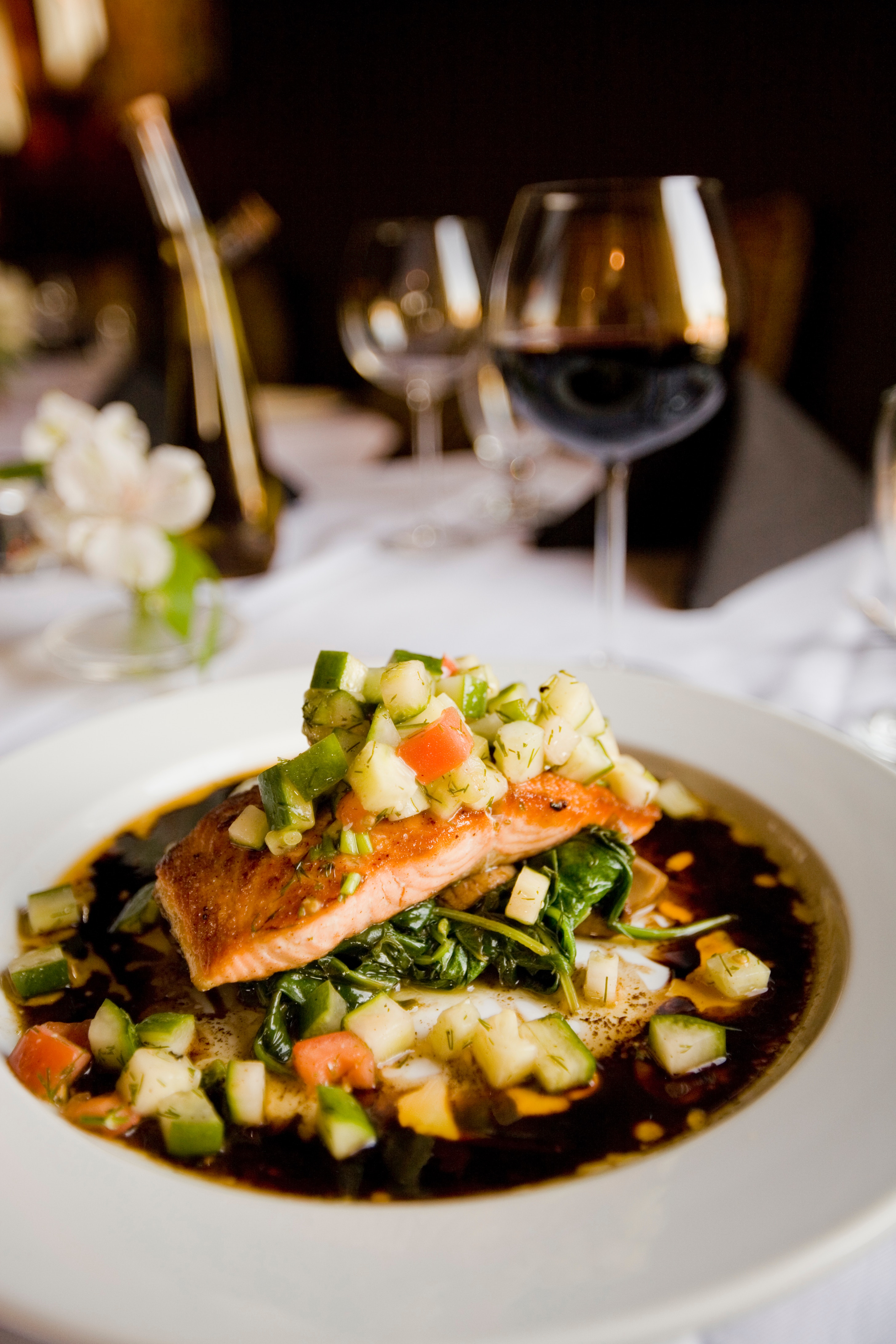 Shallow white bowl with cooked salmon on mound of cubed vegetables in brown broth, glass of red wine, bottle behind