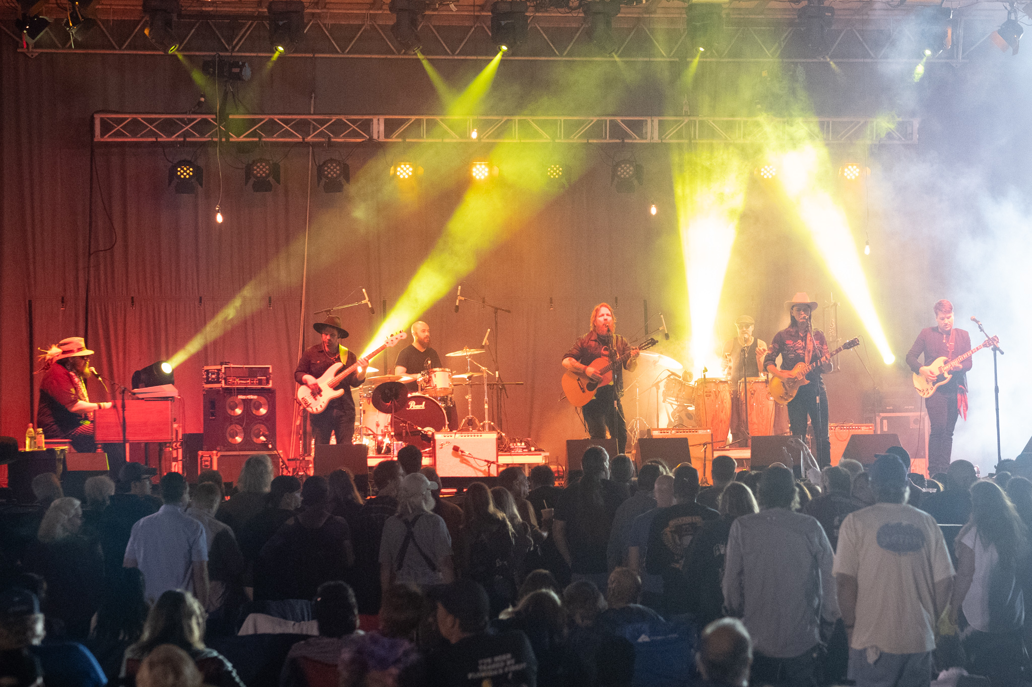 6 person band on stage with crowd in front at night 4 yellow beams of light pointing up to stage lighting above