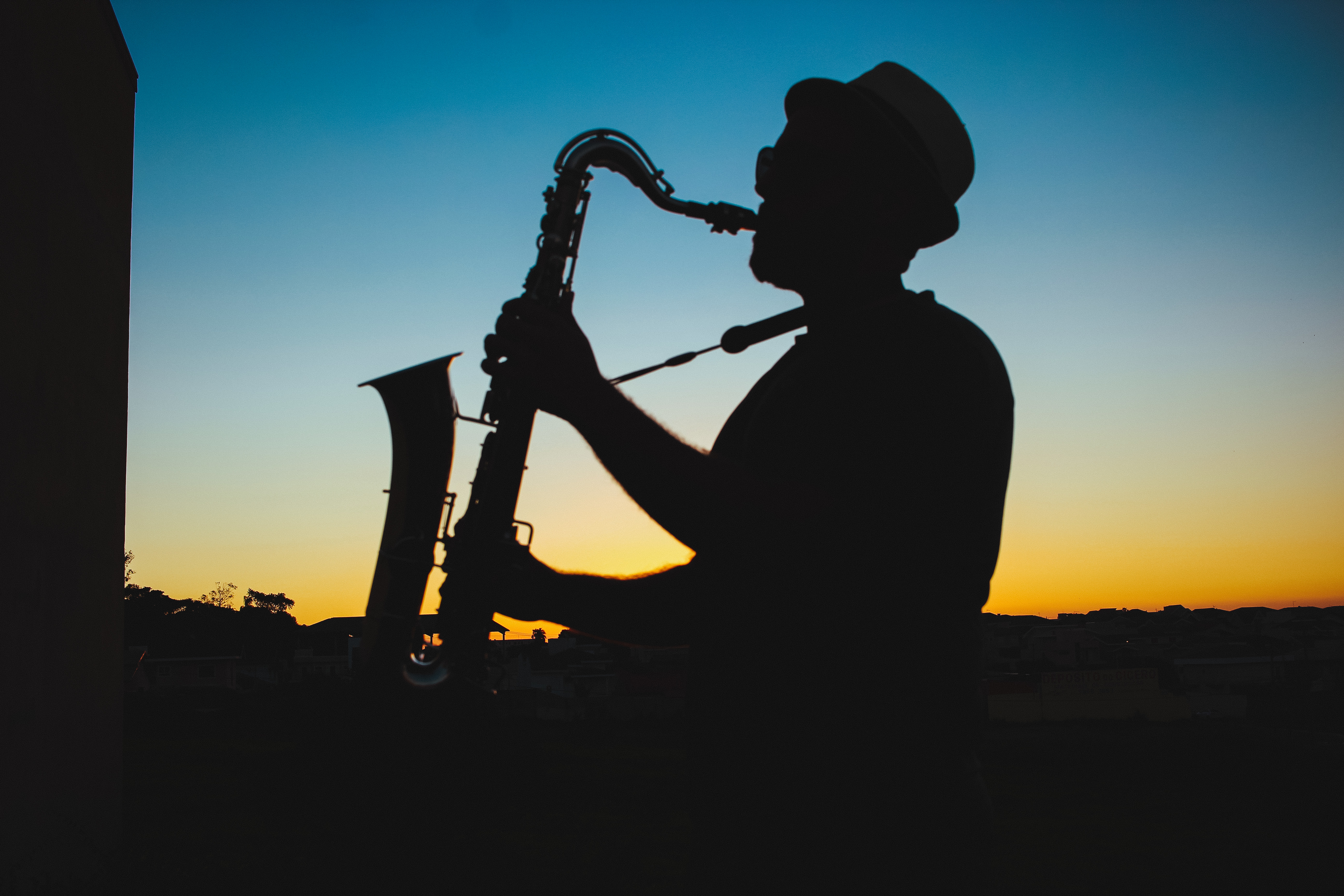 silhouette of male saxophone player in shadows wearing hat with blue and gold sunset sky behind