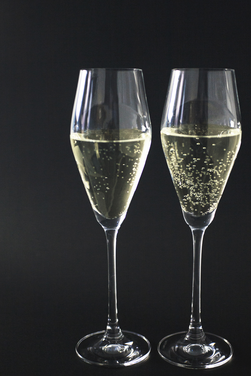 two diamond-shaped stemmed champagne glasses filled halfway with sparkling wine against black background